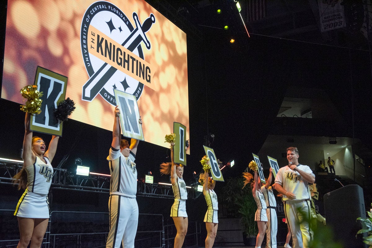 Members of cheer team hold up signs spelling KNIGHTS on stage with a Knighting banner illuminated behind them