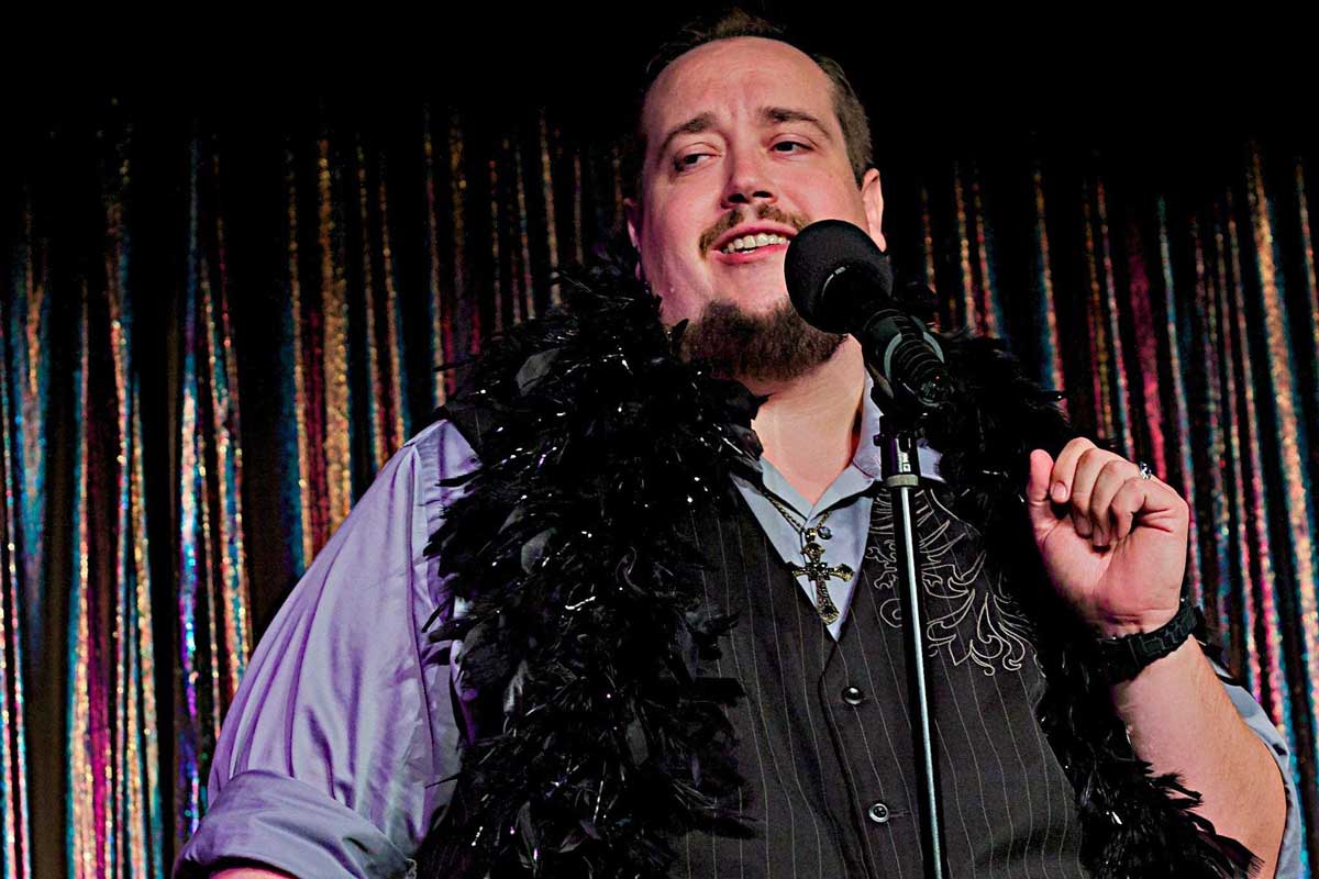 Daniel Jones with a feather boa around his neck, at a microphone