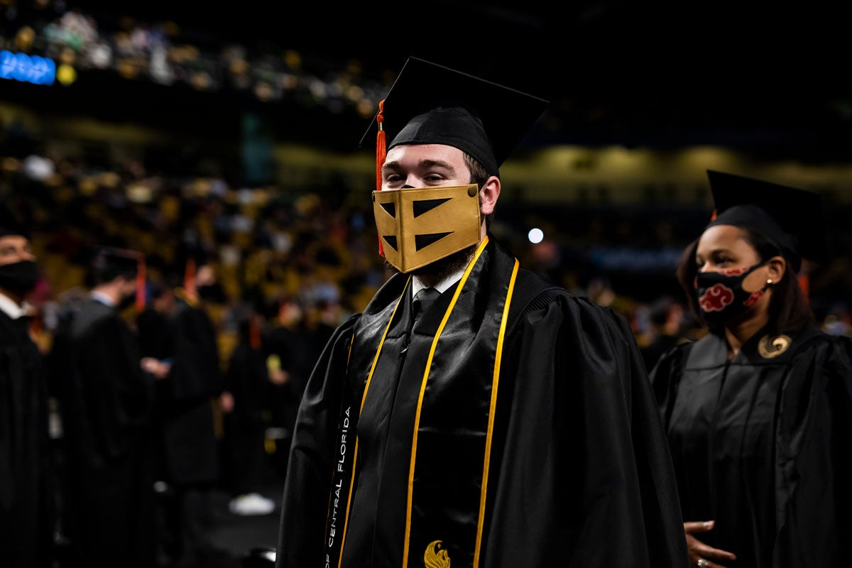 Male UCF grad in cap and gown wears a Knightro mask