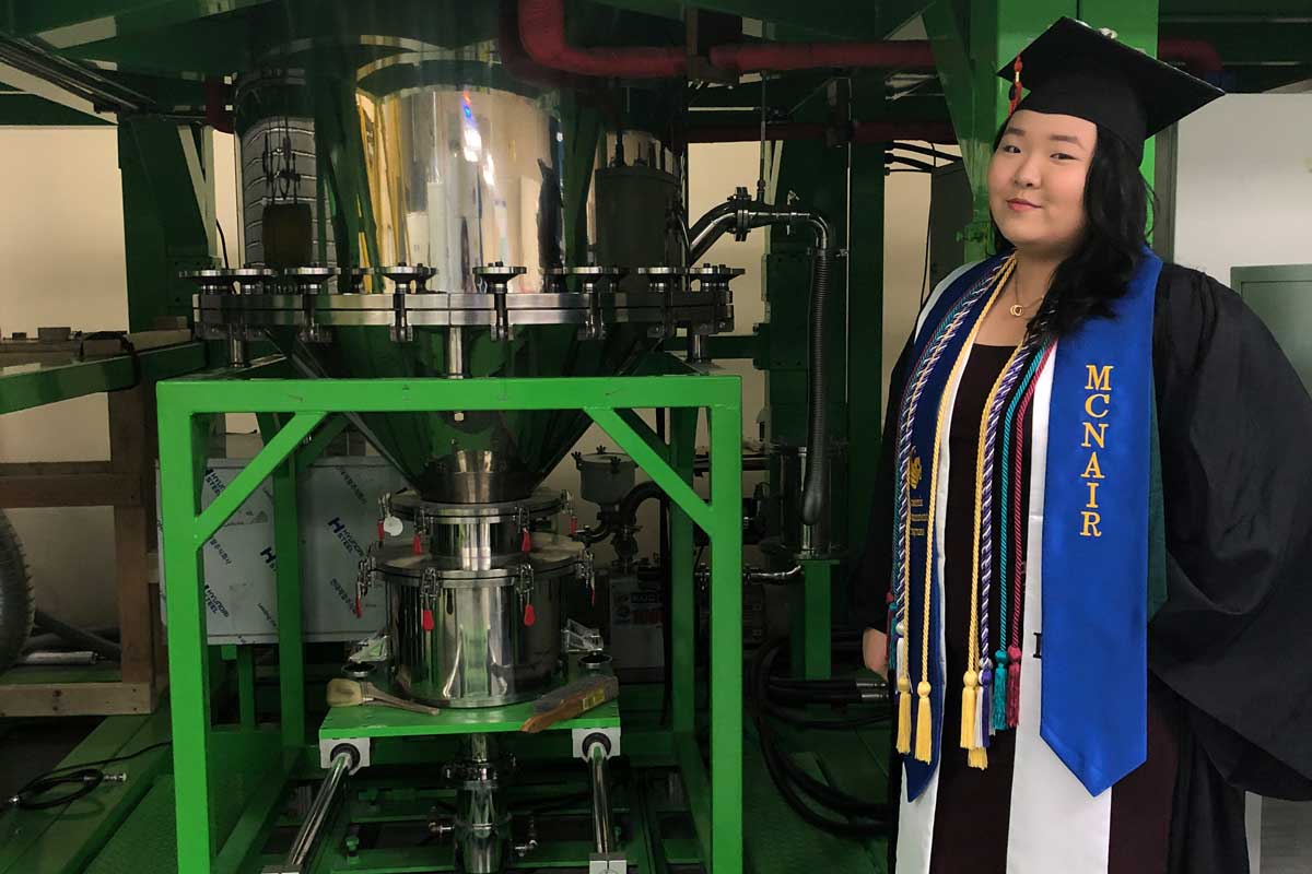 Sharon Park dressed in regalia with blue McNair stole displayed prominently, stands next to lab equipment