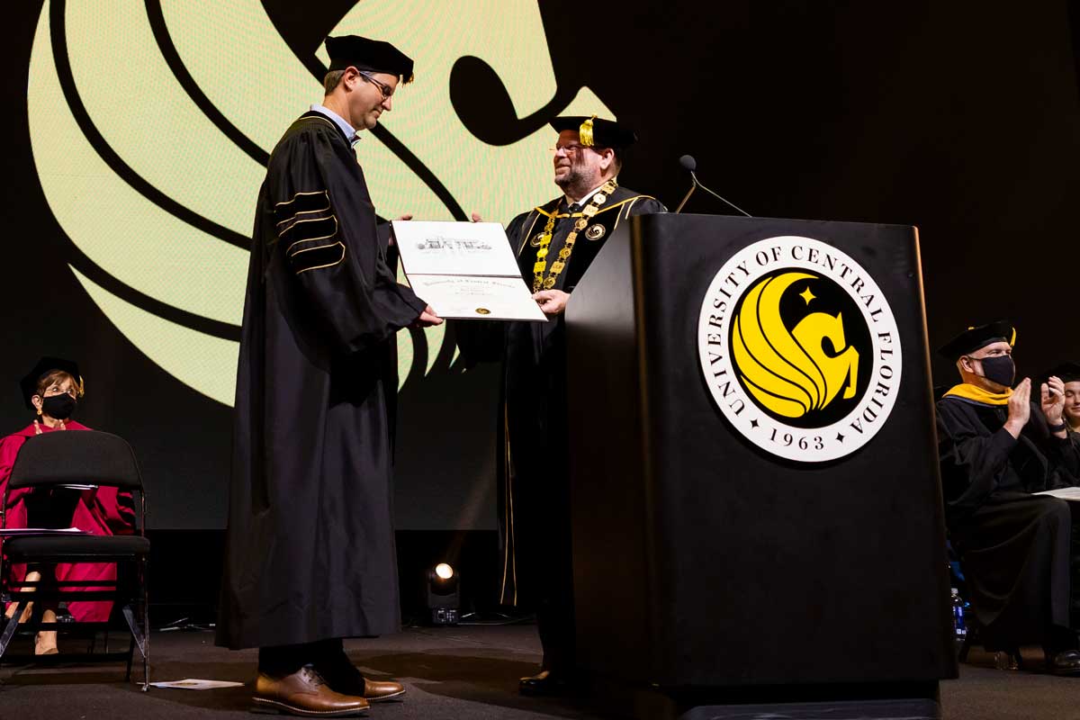 UCF President Alexander Cartwright hands Darin Edwards honorary degree on stage while both wear commencement regalia