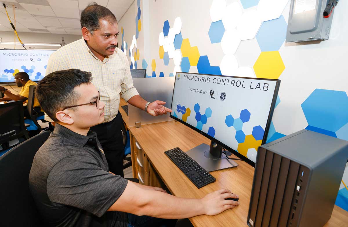 Student sits at a computer with monitor screen that reads Microgrid Control Lab as a supervisor stands next to him