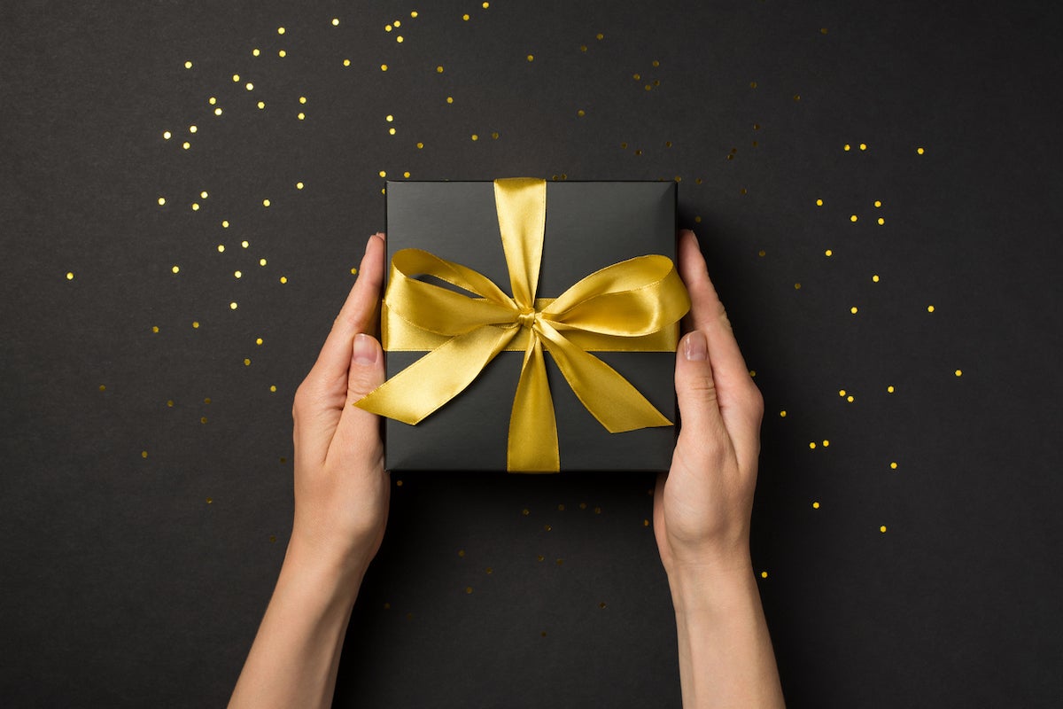 First person top view photo of hands holding black giftbox with golden ribbon bow over shiny golden sequins on isolated black background