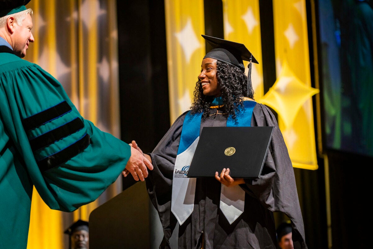 A woman wearing a grad cap and gown crosses the commencement stage while shaking a man's hand