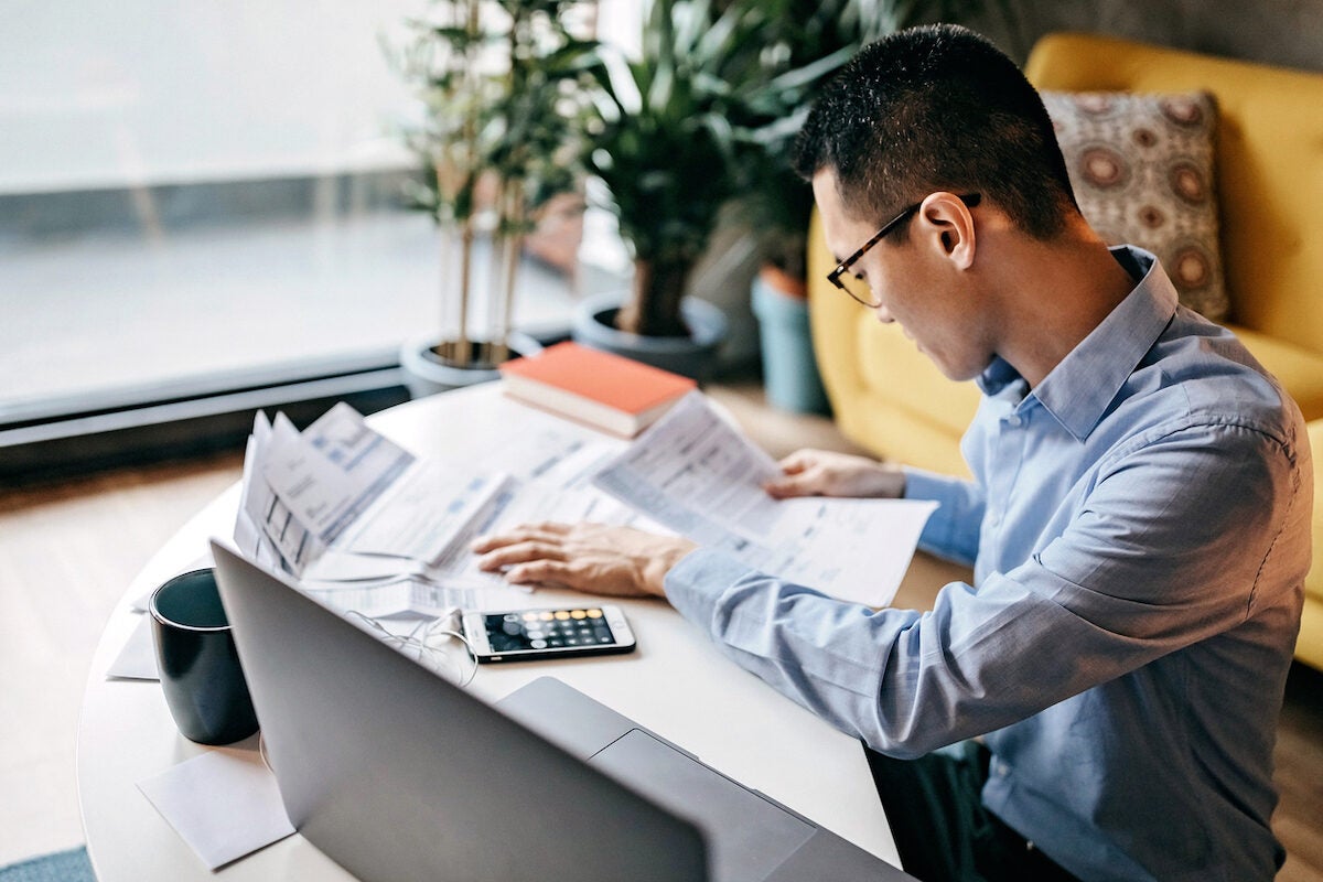 Man with eyeglasses siting on floor in the living room and using smart phone and laptop for managing home finances