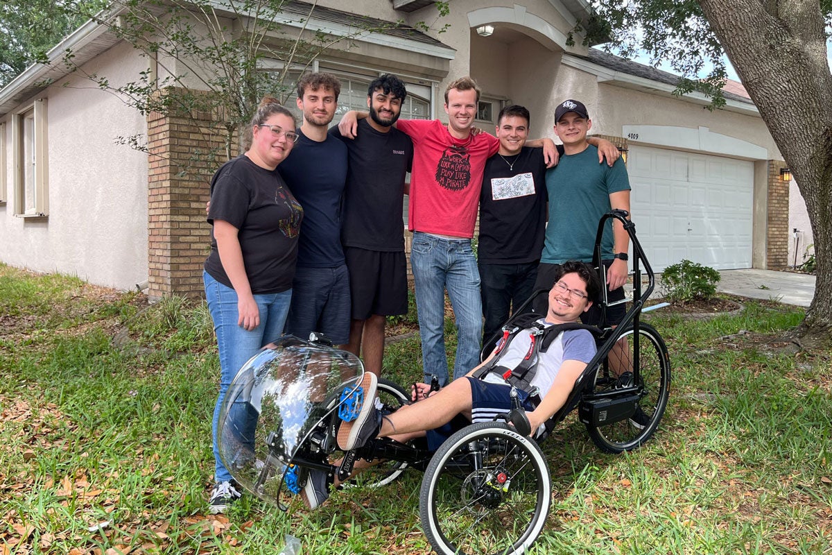 A group of engineering students pose for a photo while standing around another student sitting in their recumbent tricycle.