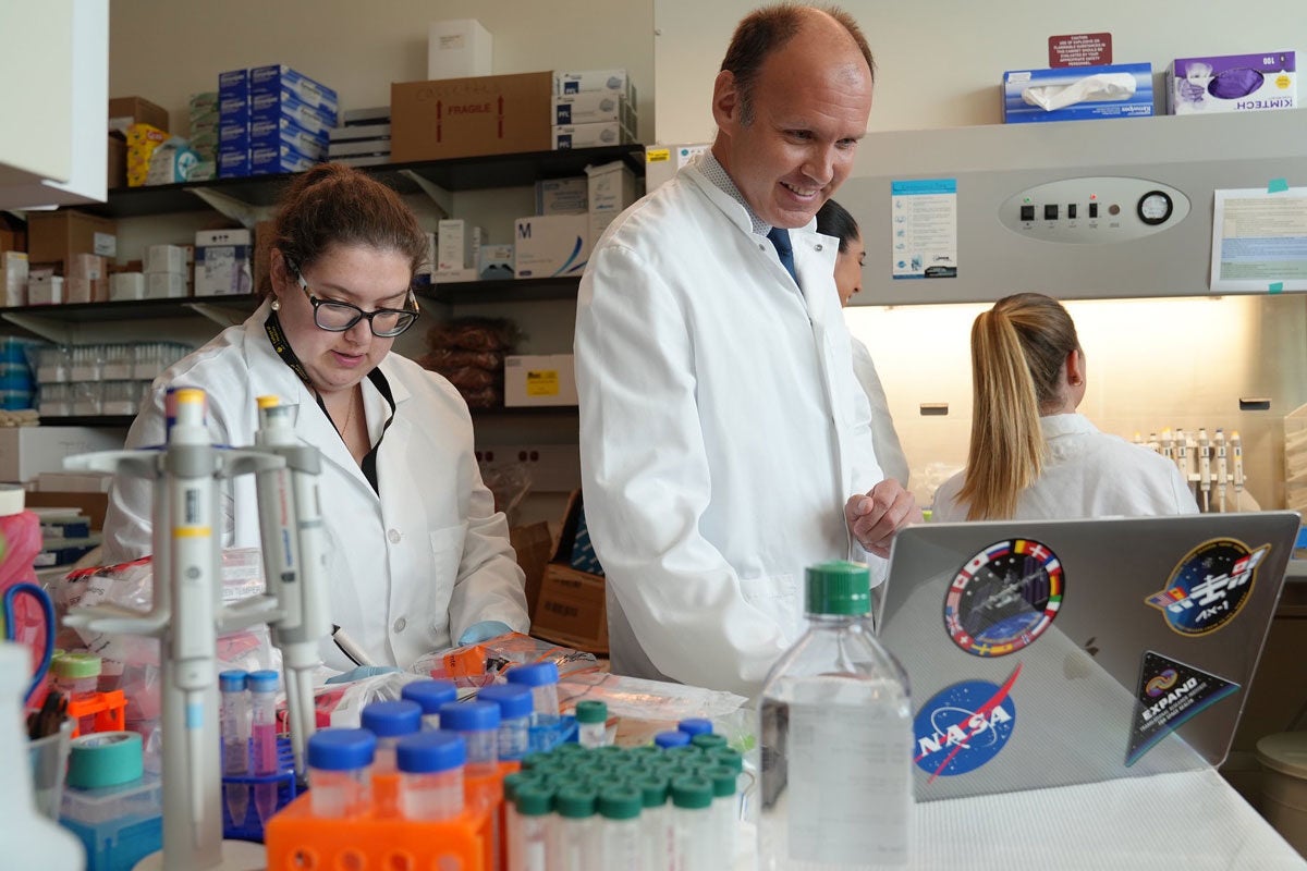 three people wearing white coats and working in a lab with a laptop with space stickers on it nearby.