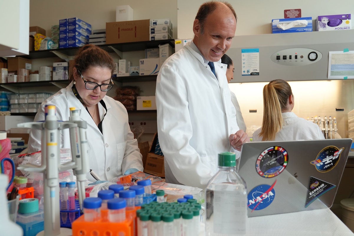 three people wearing white coats and working in a lab with a laptop with space stickers on it nearby.