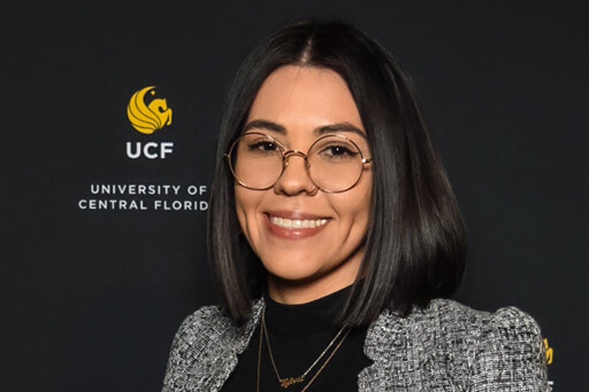 Olivia Newton poses for a photo in front of a backdrop with the UCF gold pegasus and the words"UCF. University of Central Florida."