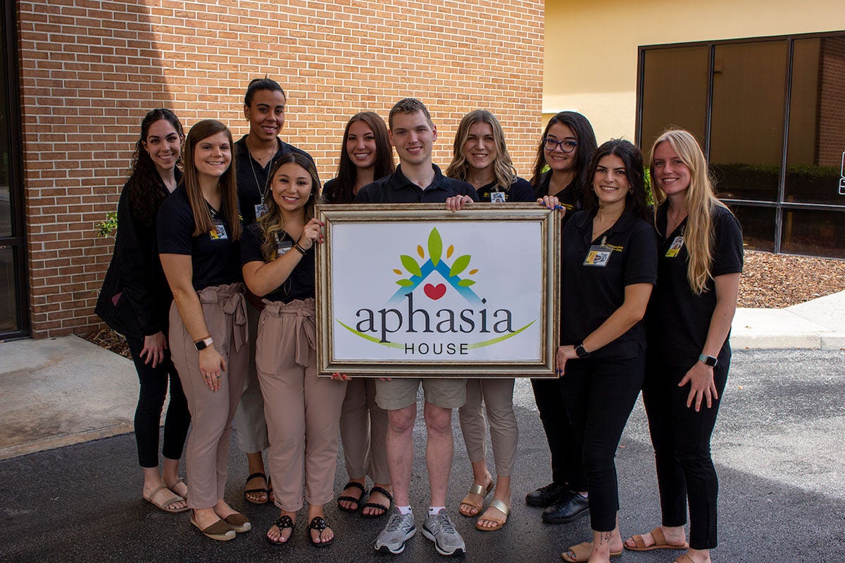 Kyle Burke and communication sciences and disorders graduate students pose for a photo while holding a UCF Aphasia House sign