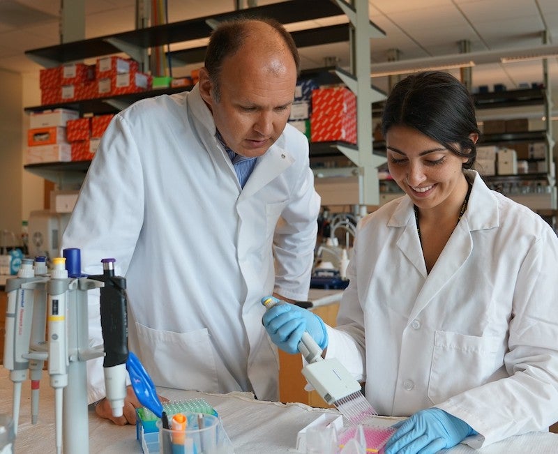 A man in white coat oversees a woman in a white coat working with lab equipment