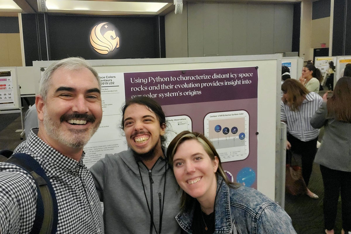 a professor and two students stand in front of a research poster display.