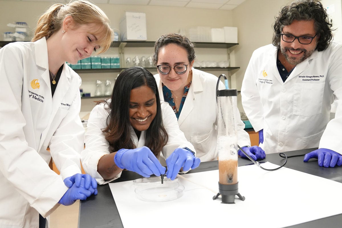 Three woman and man wear white coats and gloves while looking at a Petri dish in a lab.