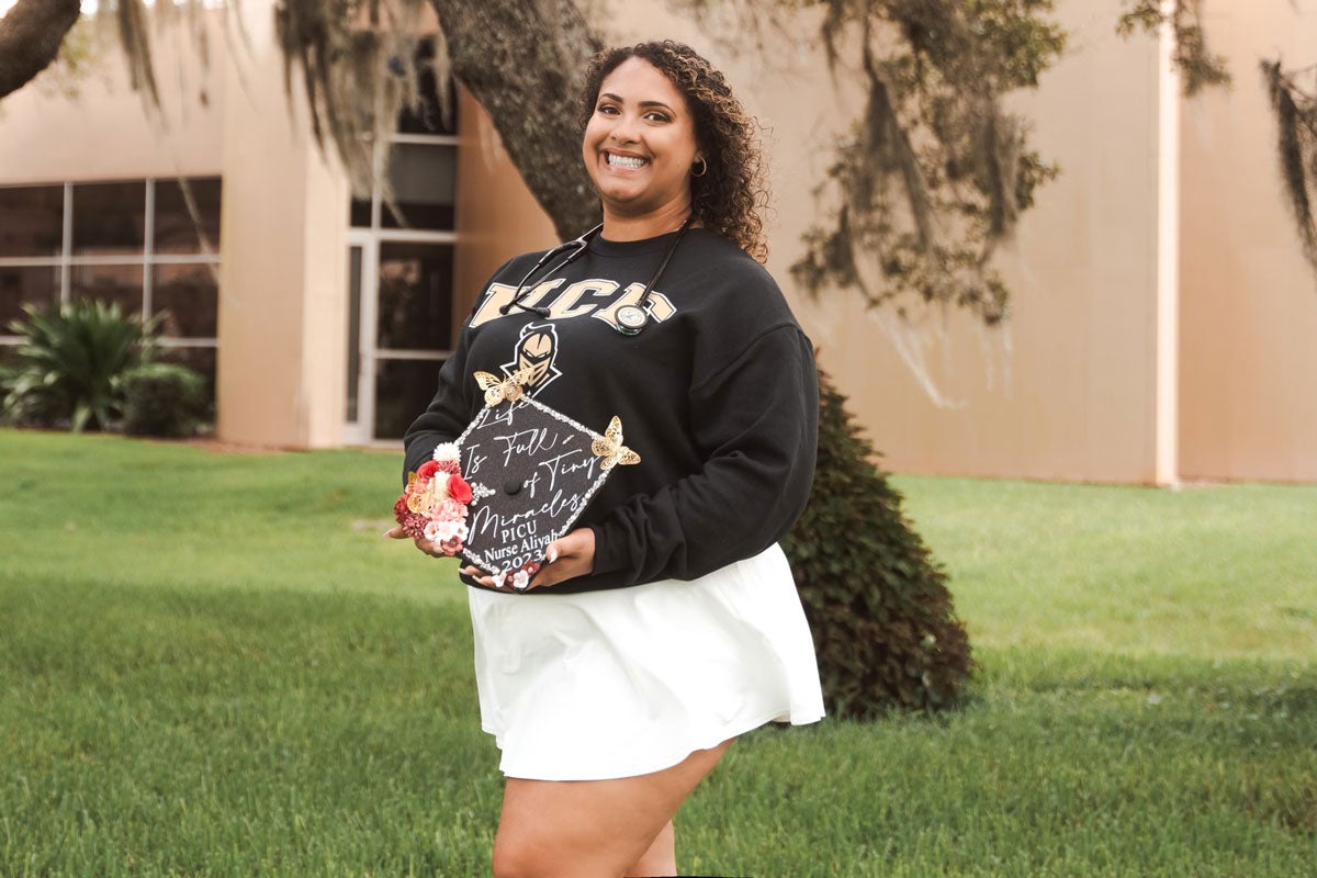 Aliyah Gonzalez holding a decorated grad cap and UCF sweater while on campus.