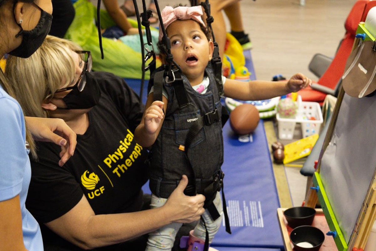 Clinical Associate Professor Jennifer Tucker uses a harness system to help a child stand during a playdate at UCF’s Rehabilitation Innovation Center.