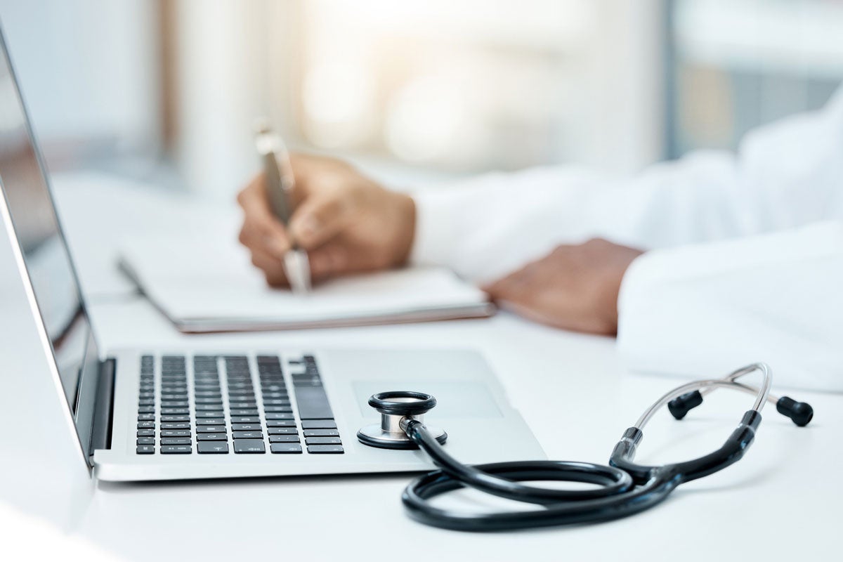 A doctor sitting at a desk writing with a laptop and stethoscope nearby