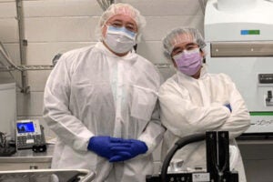 Frederick Carrick and Kiminobu Sugaya dressed in white coats, gloves, masks and hairnets in a lab. 