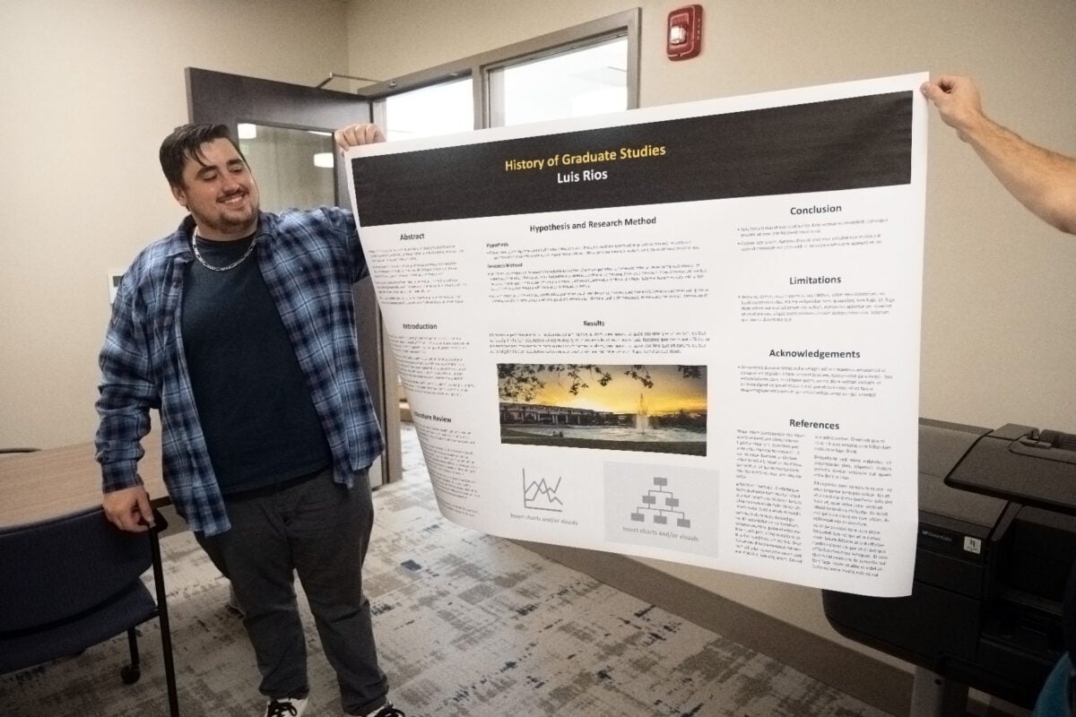male in blue checkered shirt holding one end of a poster usually used to explain research.