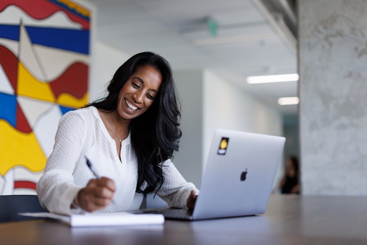 A woman smiling and writing on a notepad while sitting in front of an open laptop with a UCF sticker on it