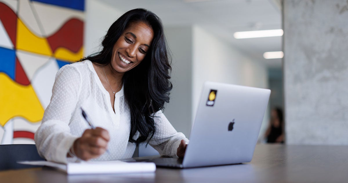 A woman smiling and writing on a notepad while sitting in front of an open laptop with a UCF sticker on it