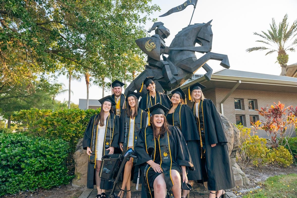 A group of students wearing graduation caps and gowns, standing in front of a Knight statue.