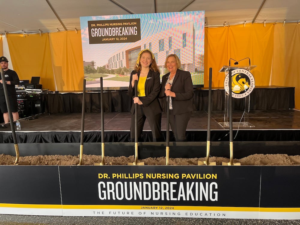 Rebekah May and College of Nursing Dean Mary Lou Sole digging shovels into dirt with the word "Groundbreaking" on a banner in front of them