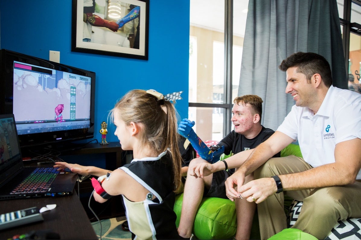 A man and a child with a bionic limb watches another child with a bionic limb play a video game
