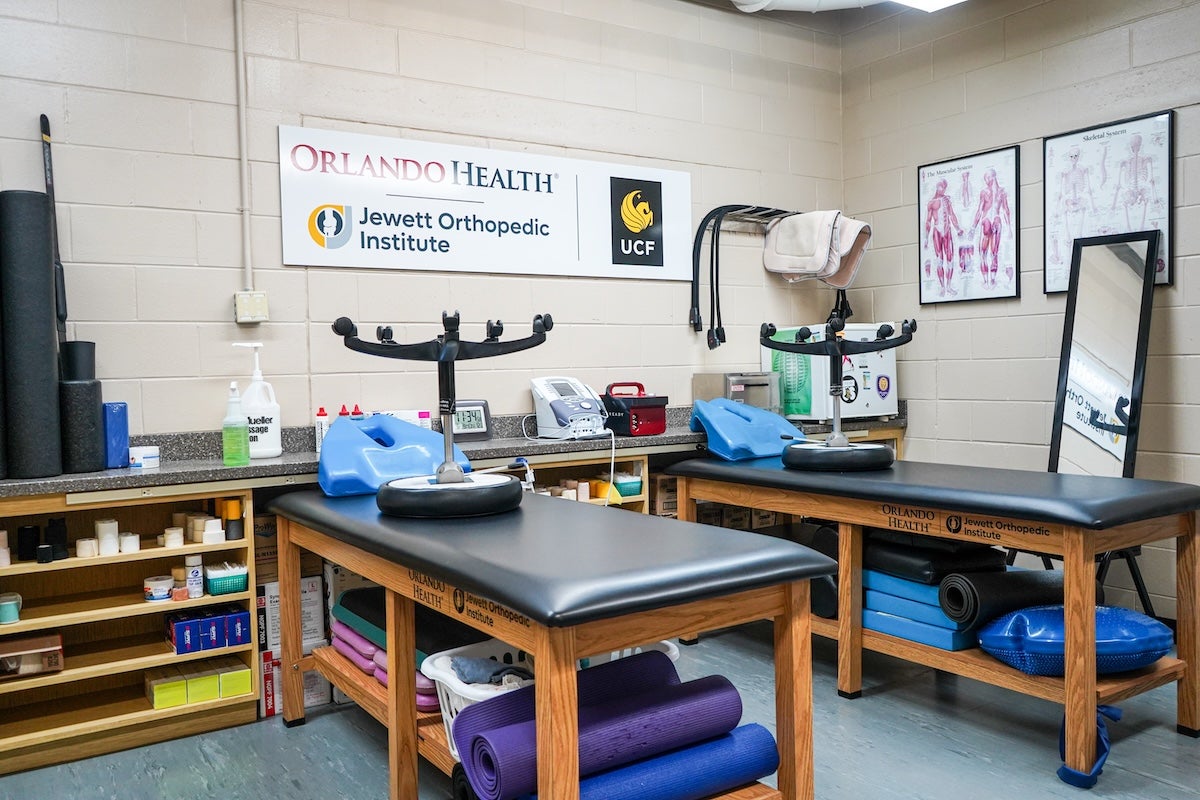 The image shows a physical therapy or rehabilitation room with two black padded treatment tables. Each table has a blue bolster and a black armrest device on top. The room has shelves with various medical supplies, including rolls of tape, bottles of lotion, and exercise mats. On the wall above the counter is a sign that reads 'Orlando Health Jewett Orthopedic Institute' with the UCF logo next to it. There are anatomical charts on another wall and a full-length mirror leaning against the wall.