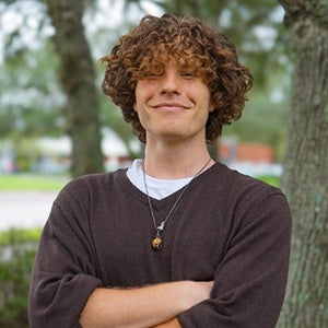 UCF biomedical science student Ethan Bell stands outside with arms crossed, smiling confidently in front of trees.