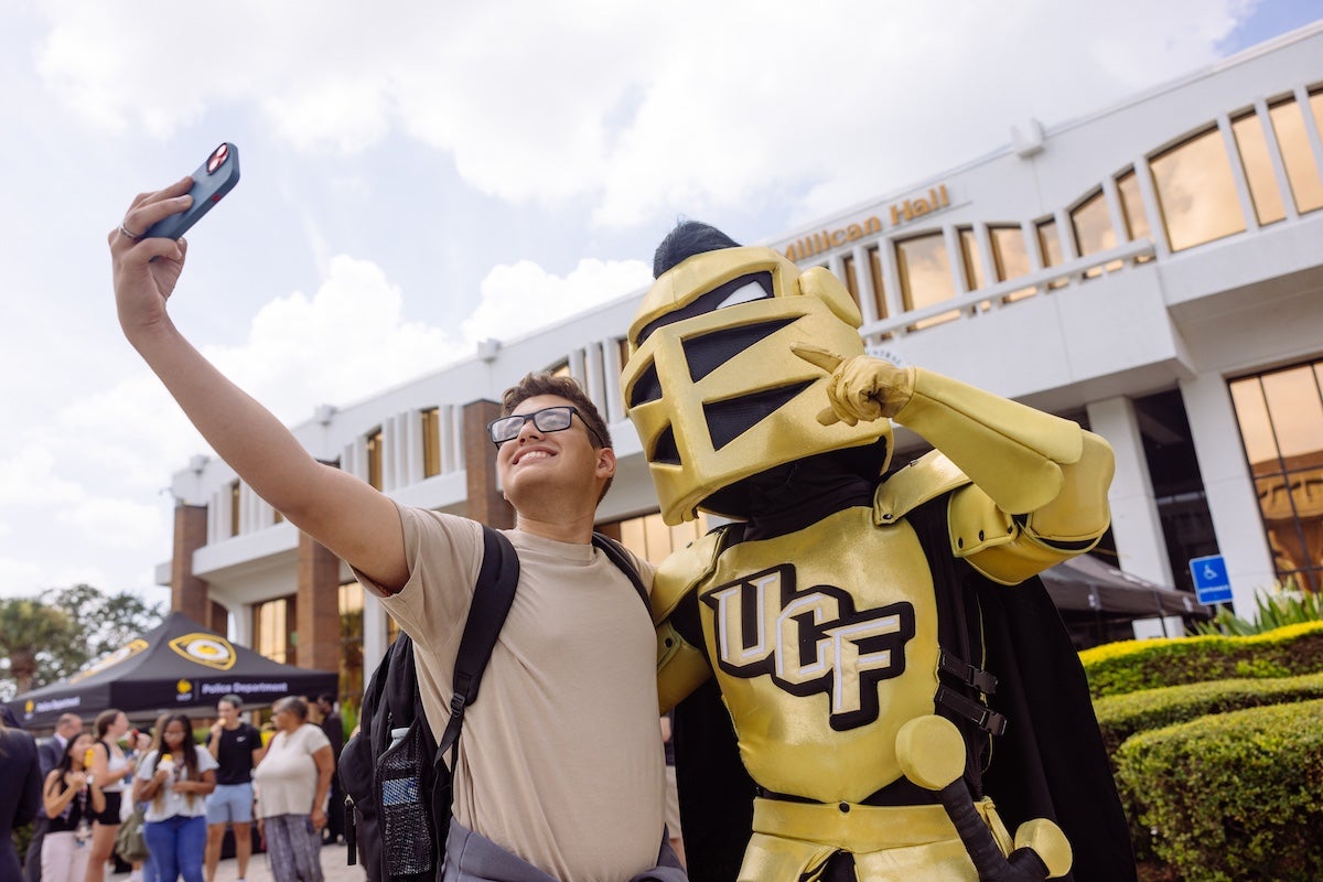A UCF student taking a selfie with Knightro in front of Millican Hall at the main campus.