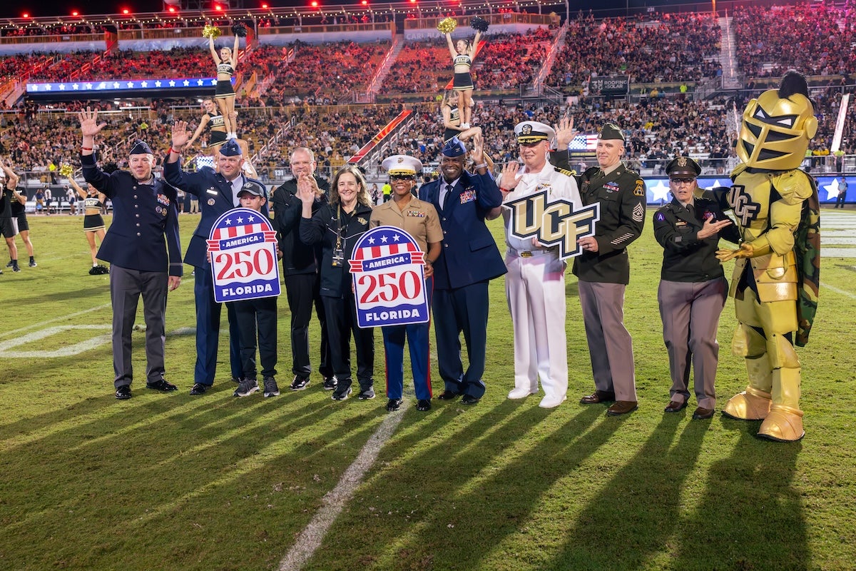 Knightro stands with members of the military dressed in uniform on the football field at Acrisure Bounce House