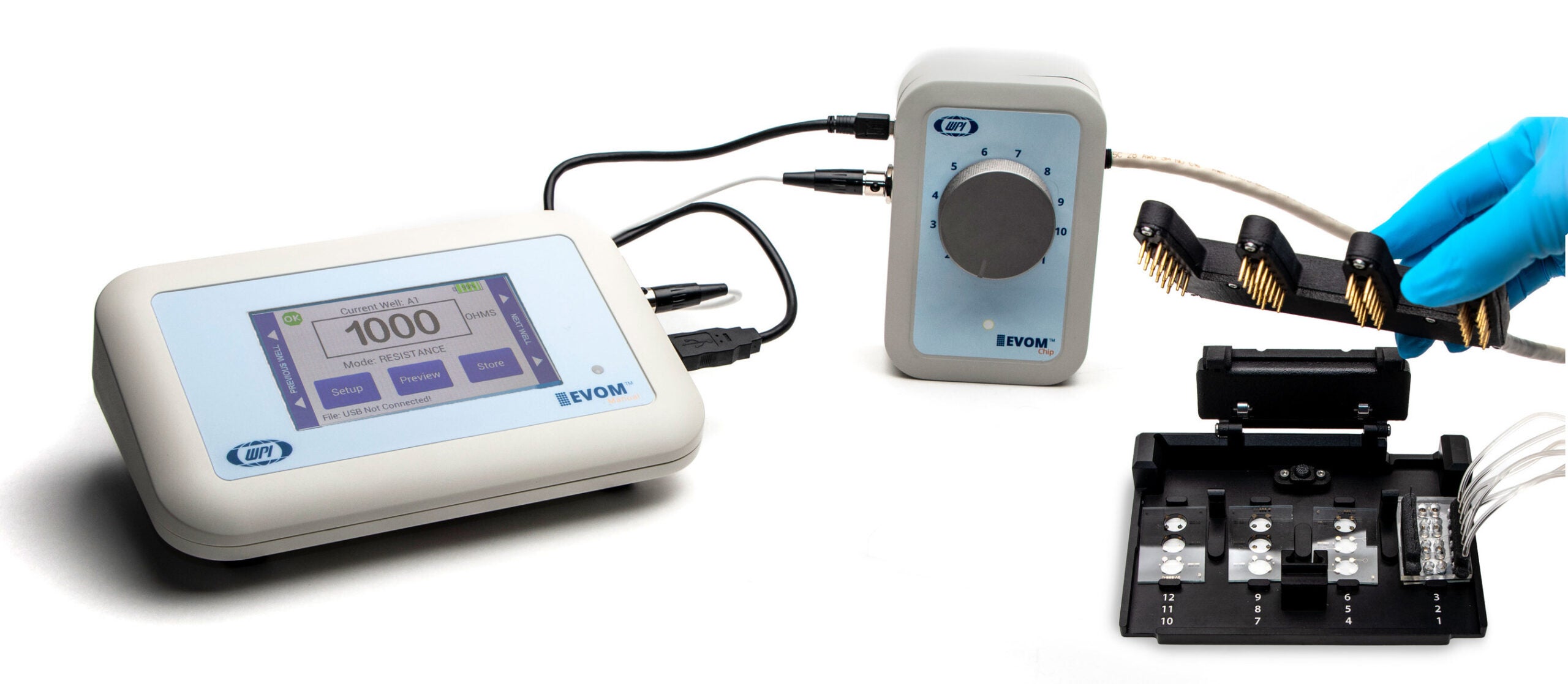 Laboratory setup featuring WPI's EVOM3 system with a touchscreen device displaying '1000' and options for 'Setup,' 'Measure,' and 'Store.' The system is connected to a central unit with a numbered dial and an open chip platform labeled 1 to 12. A gloved hand holds a gold-plated electrode array above the platform, which contains small wells for biological sample testing.