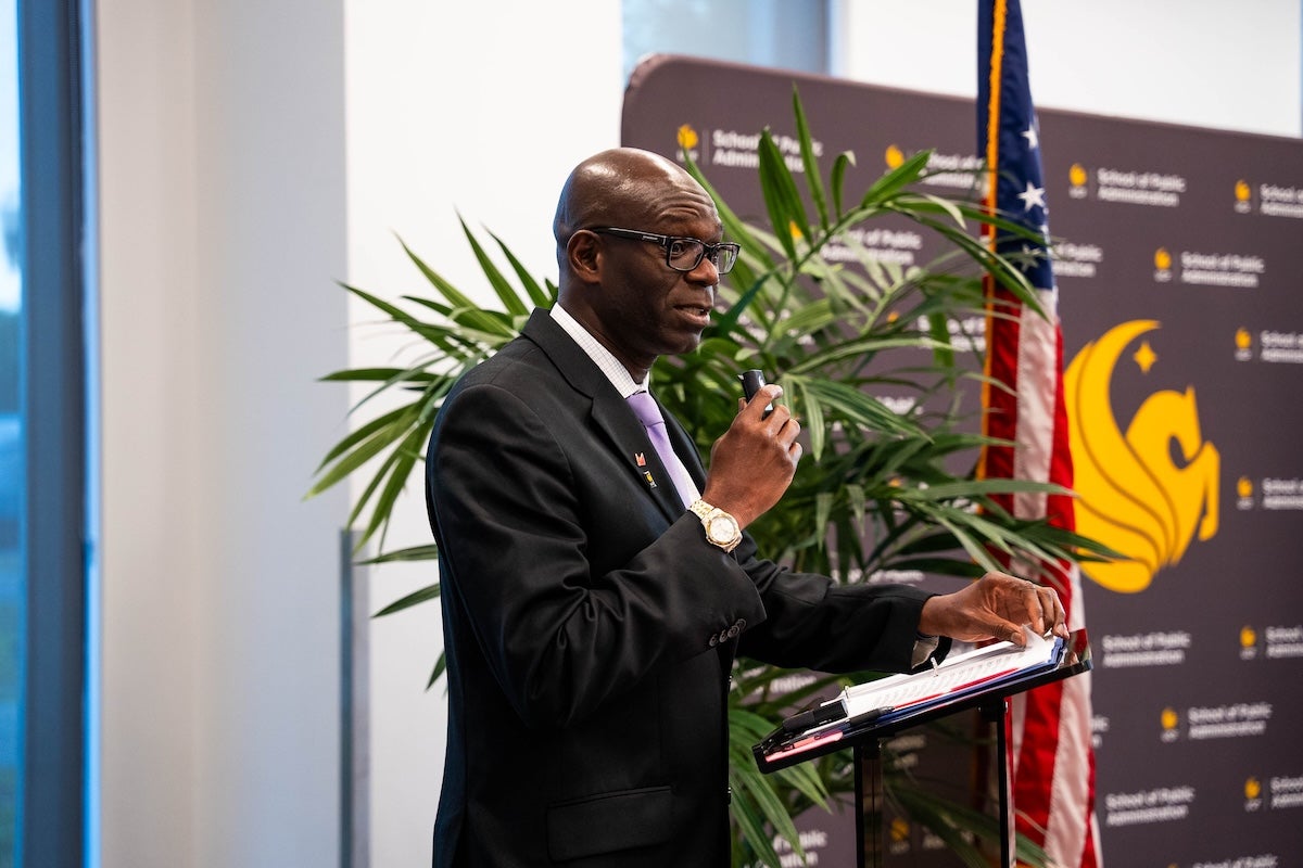 Abdul-Akeem Sadiq, wearing a suit and holding a microphone, stands in front of a podium speaking in a classroom at UCF Downtown.