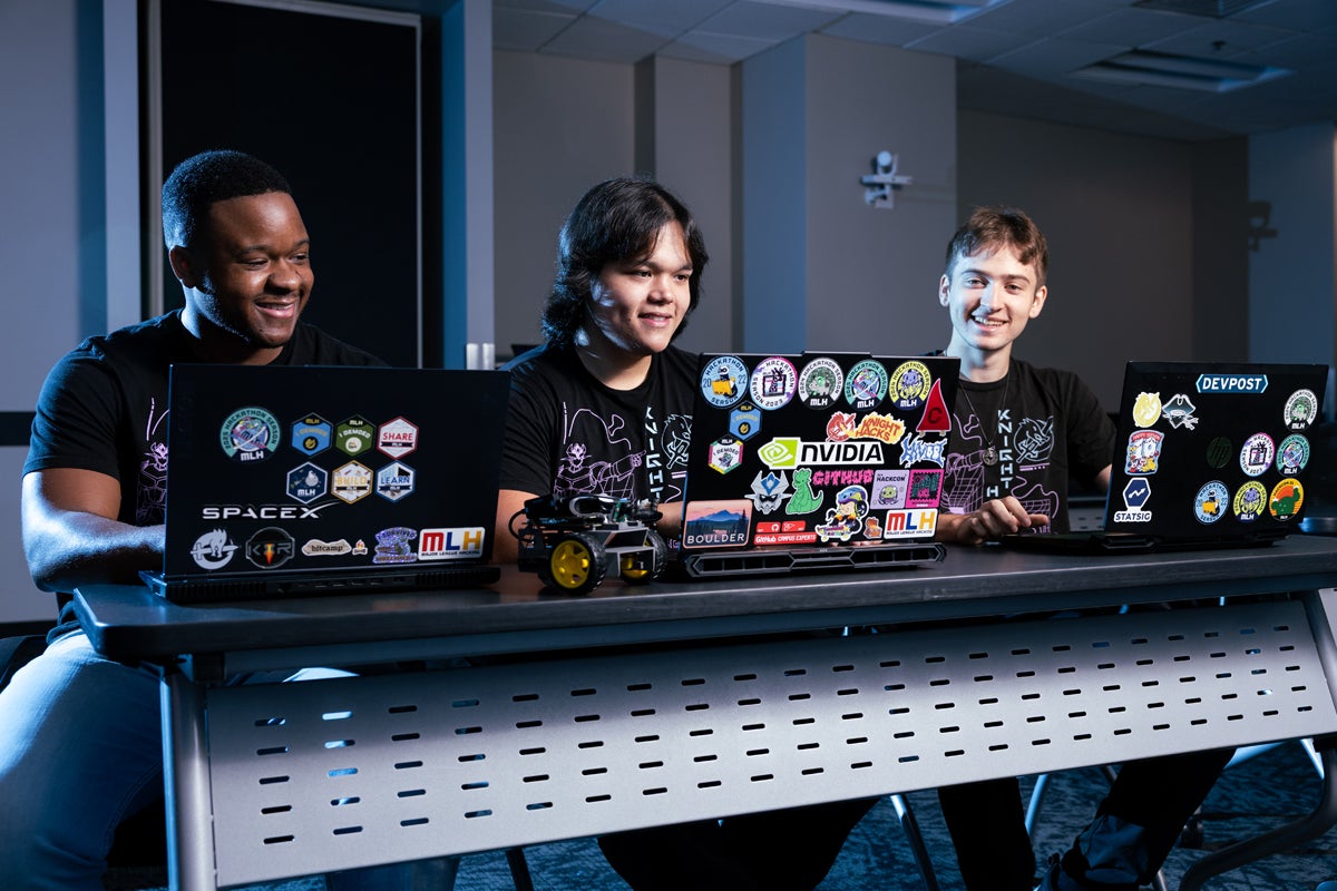 Three individuals seated at a table, each working on laptops decorated with tech-related stickers including SpaceX, NVIDIA, and Devpost