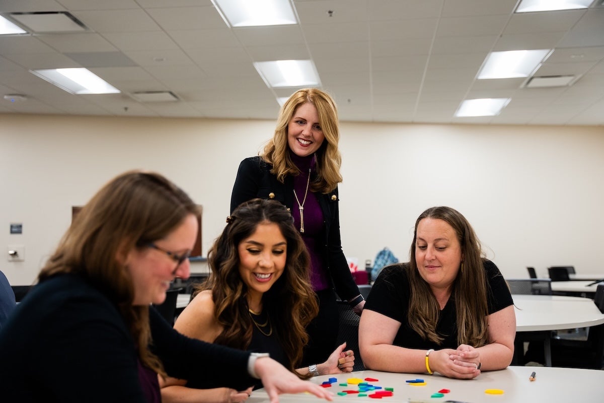 Classroom mathematics teachers sit at a table engaging in mathematics discourse while Sarah Bush facilitates in the background.