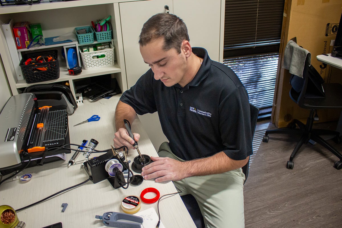 A young man in a black polo shirt and khaki pants sits at a desk holding a tool, modifying equipment.