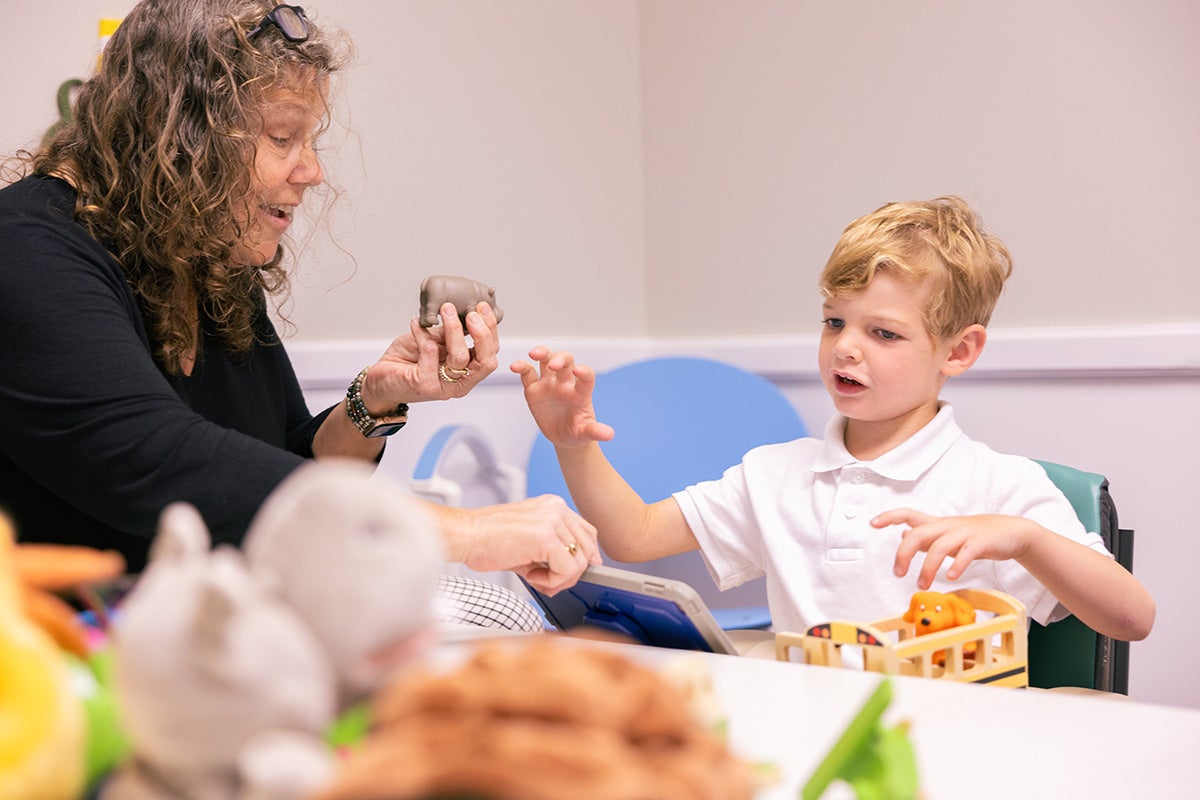 Nancy Harrington sits at a table with a young, blonde boy in a white collar shirt, ready to hand him a toy Hippo.