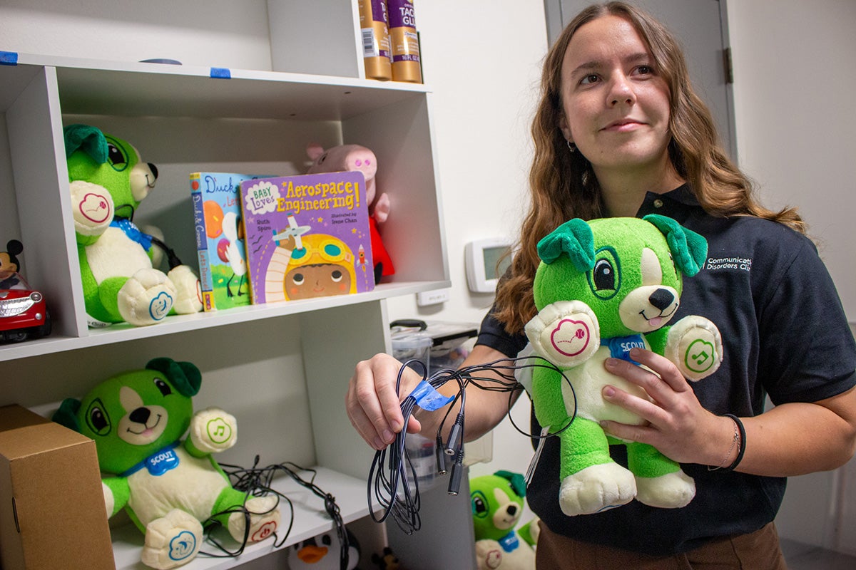 A brunette young woman holds a green stuffed animal dog with wires in a storage closet.