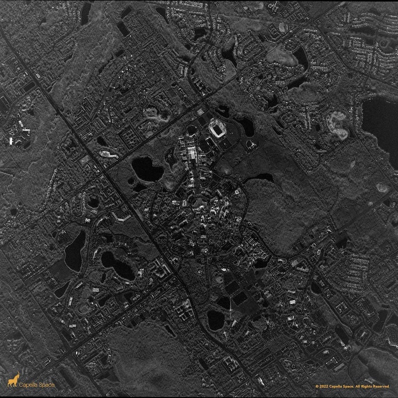 aerial satellite image of the University of Central Florida (UCF) campus and surrounding area. The image shows a dense cluster of campus buildings arranged in a circular layout at the center, with a large stadium visible near the top. Roads intersect diagonally and horizontally, connecting residential neighborhoods and commercial areas. Several lakes and green spaces are scattered throughout the scene, captured in high-resolution black-and-white radar imagery.