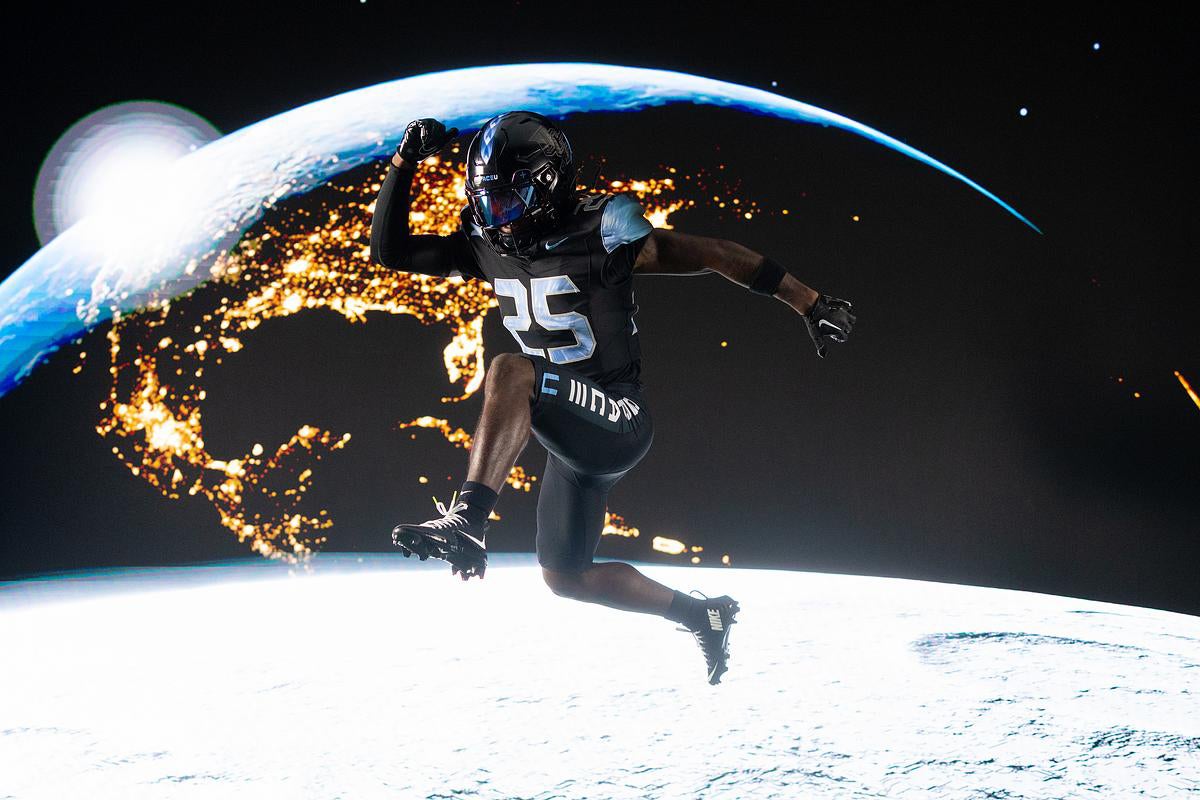 A football player wearing a black uniform with the number 25 and a black helmet leaps in midair against a dramatic backdrop of Earth viewed from space. The Earth shows illuminated city lights across North America and a glowing horizon, creating a futuristic, space-themed setting.