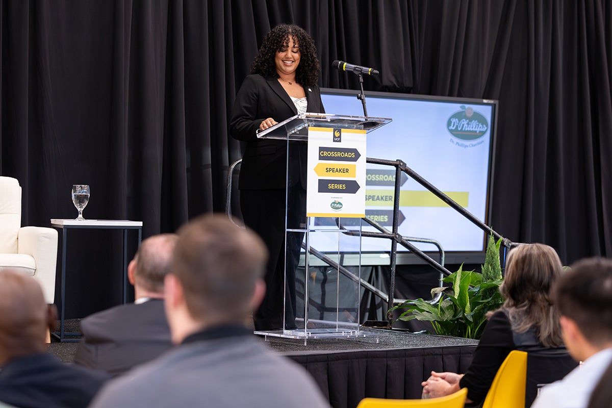Black woman wearing a black business suit stands at a podium with "Crossroads Speaker Series" signage.