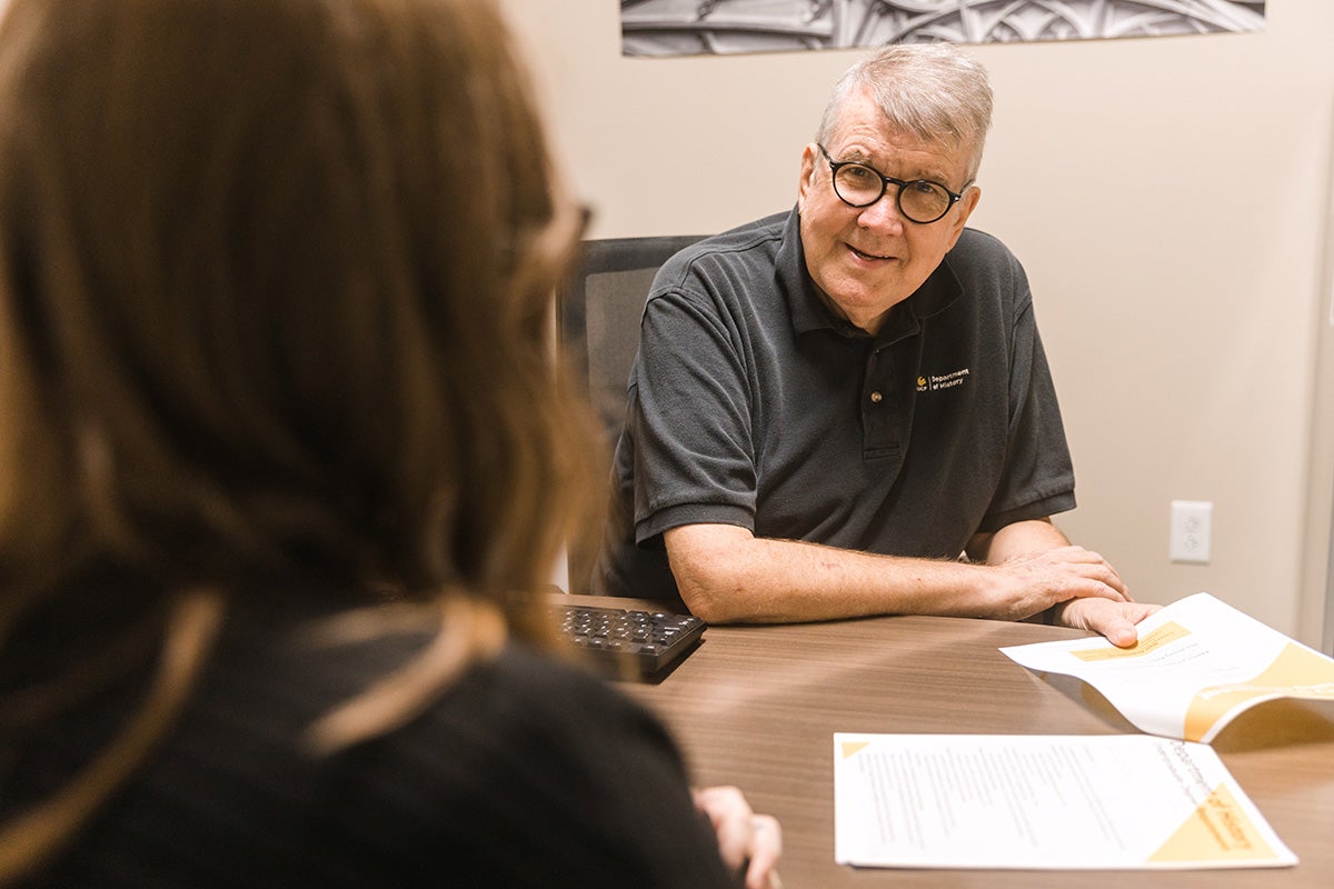 Jim Clark, man with gray hair and black glasses wearing gray polo shirt, holds a paper at his desk