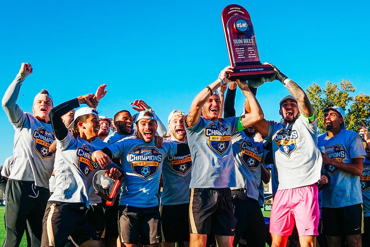UCF men's soccer team, wearing gray Sun Belt Champions T Shirts, hold brown trophy in air against clear blue sky.