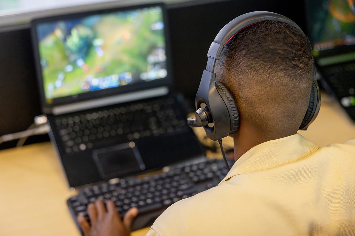 Black male sits at a desk while wearing black headphones, typing on a keyboard and focusing on a monitor.