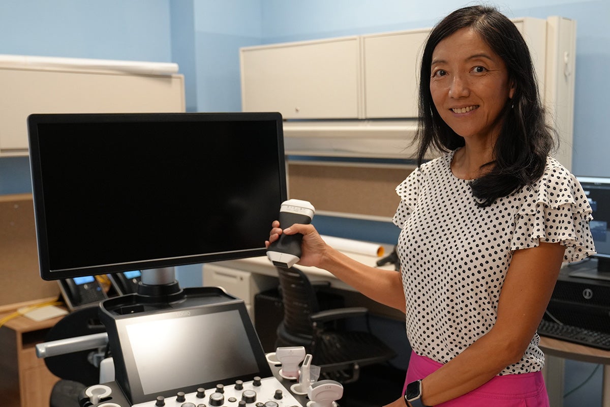Laura Brattain, dressed in white and black polka dot ruffle blouse and pink pants poses in front of computer monitor and ultrasound screen.