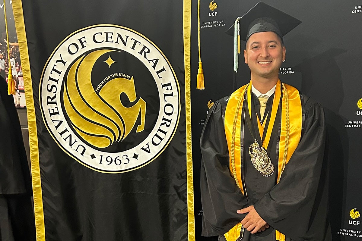 Male in black and gold cap and gown stands next to banner with UCF seal