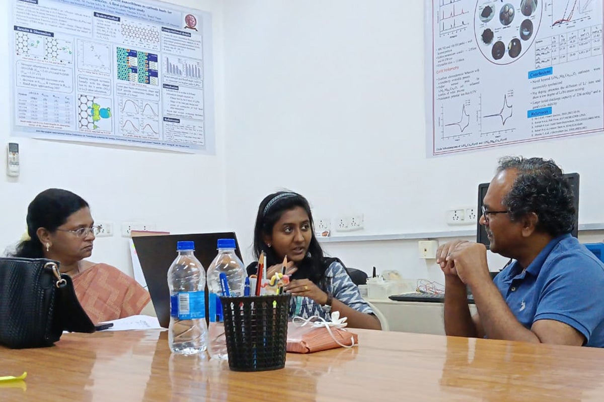 Two women and one man sit at a wooden desk with large scientific posters on walls behind them