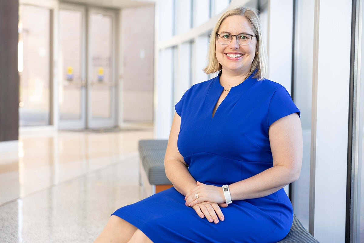 Blonde woman wearing glasses and blue dress sits on a bench near a window