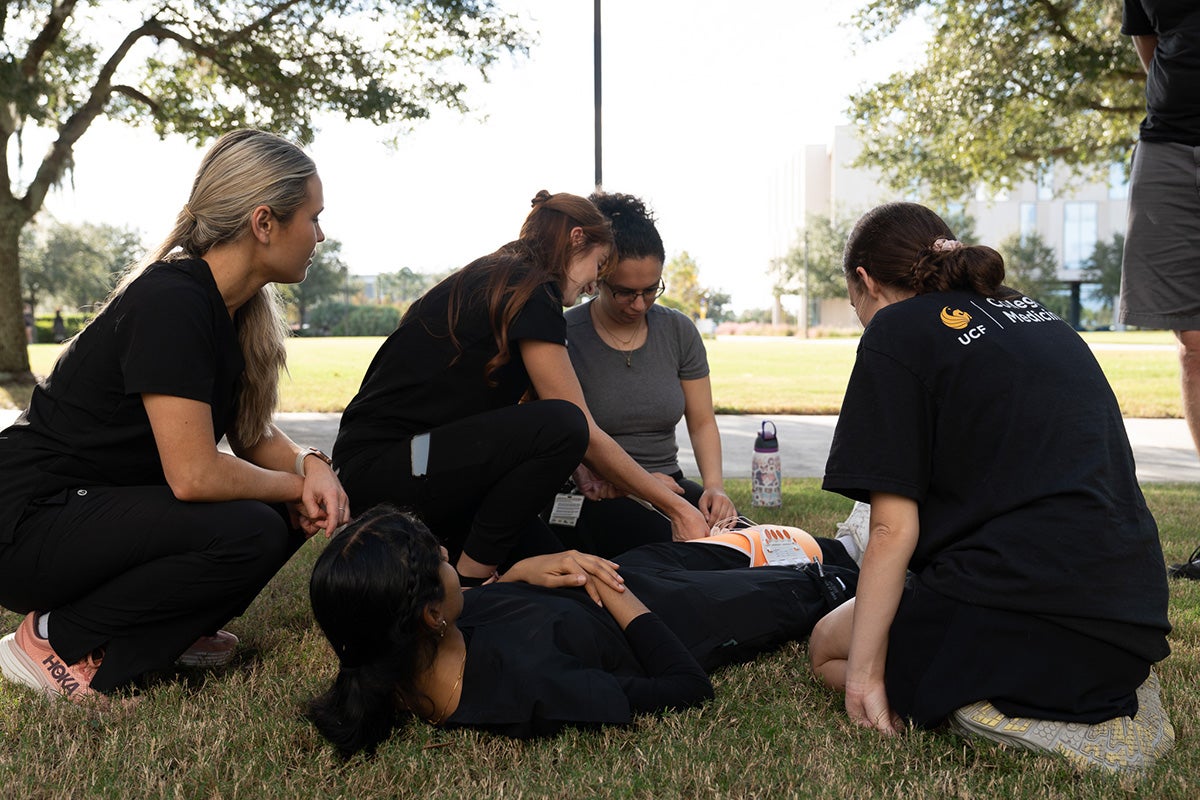 group of three medical students practice bracing a patient's leg while she lays on the grass