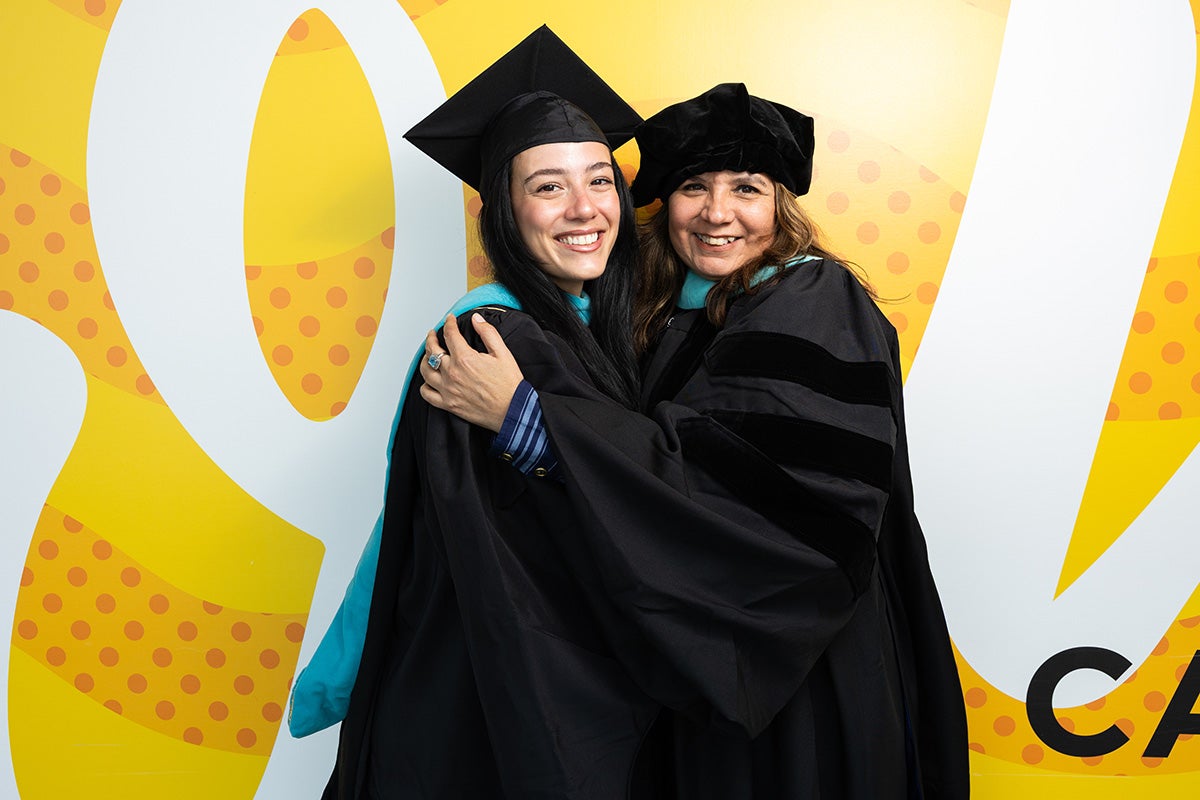 Ketty Dones and Maritza Concha, wearing traditional commencement robes, embrace in front of gold wall